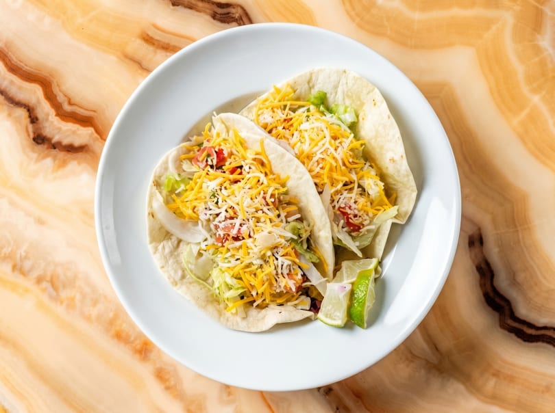 Two soft tacos with shredded cheese, lettuce, and tomatoes, served with lime wedges on a white plate.