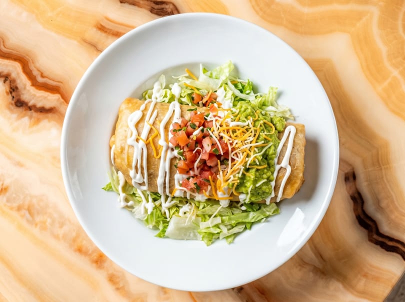 A golden-brown chimichanga topped with sour cream, pico de gallo, guacamole, and shredded cheese.