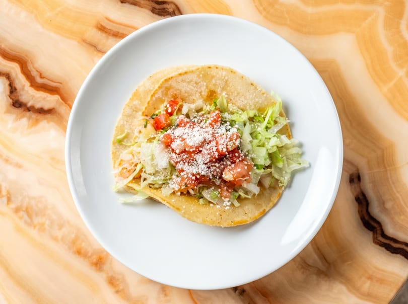 A single taco on a white plate, topped with lettuce, diced tomatoes, and cotija cheese.